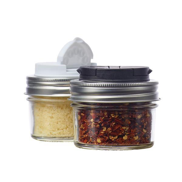 A pair of white and black plastic Jarware Mason Jar Spice shaker lids containing parmesan cheese and chilli flakes on a white background. 